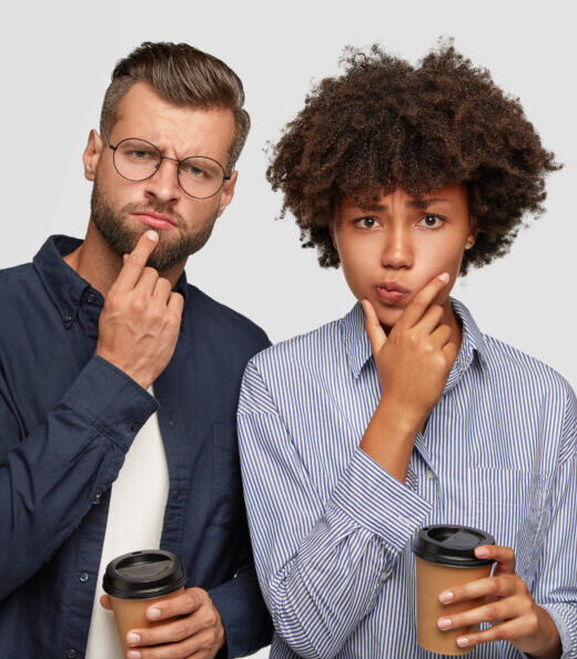 Shot of thoughtful puzzled multiethnic young couple hold chins, try to solve problem, drink aromatic takeaway coffee, stand closely against white background with copy space for your advertisement